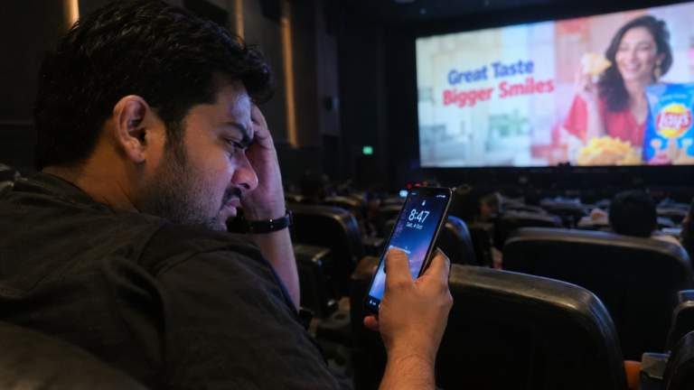 An Indian consumer checking the time on his phone in a dark cinema hall while advertisements play on the screen.